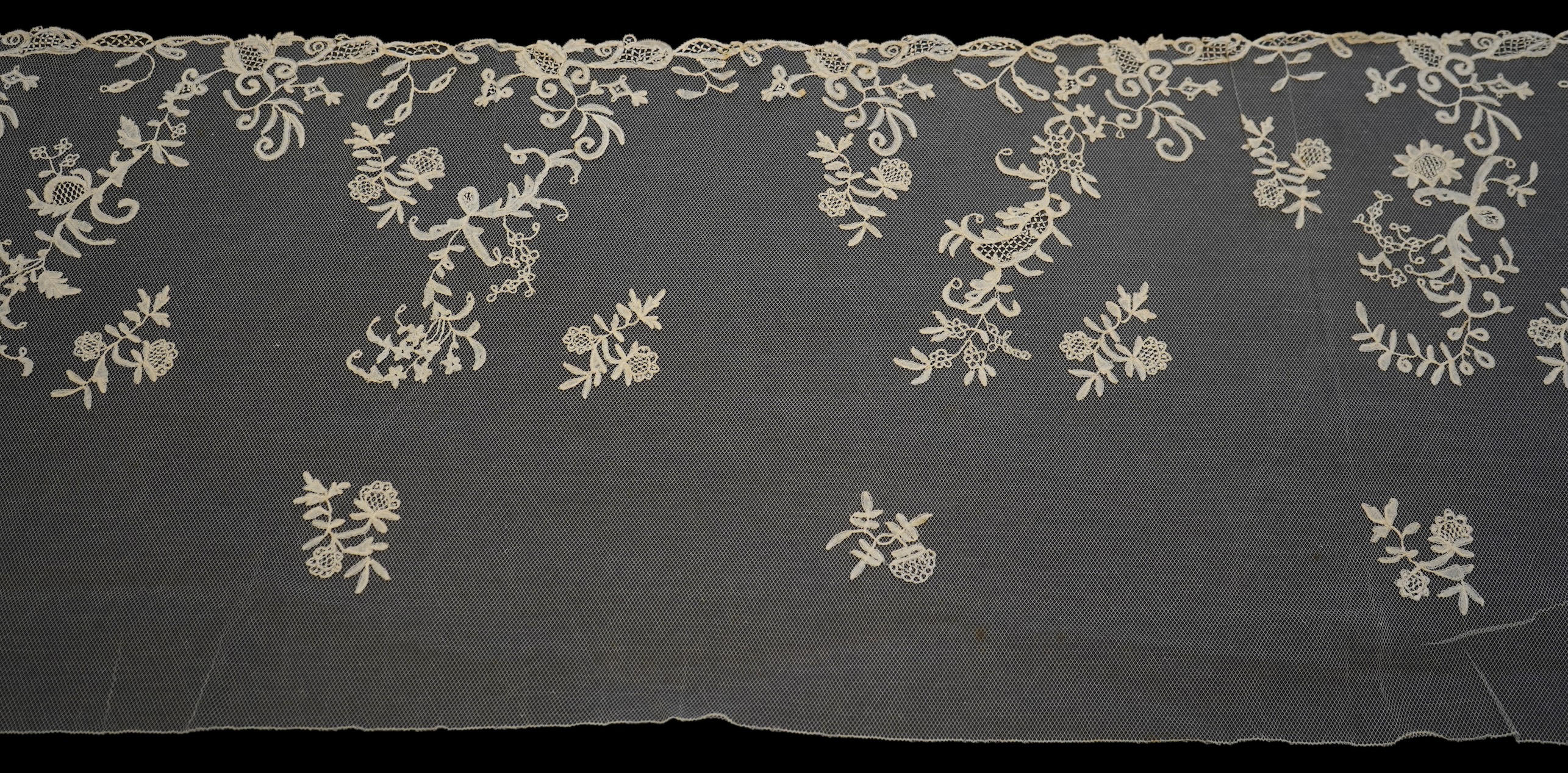 A 19th century five yard flounce of Brussels bobbin appliqué lace applied onto fine machine net, worked in a trailing floral design with an ornate border and sprig motifs, 457.5 cm long x 36cm deep. Condition - possibly